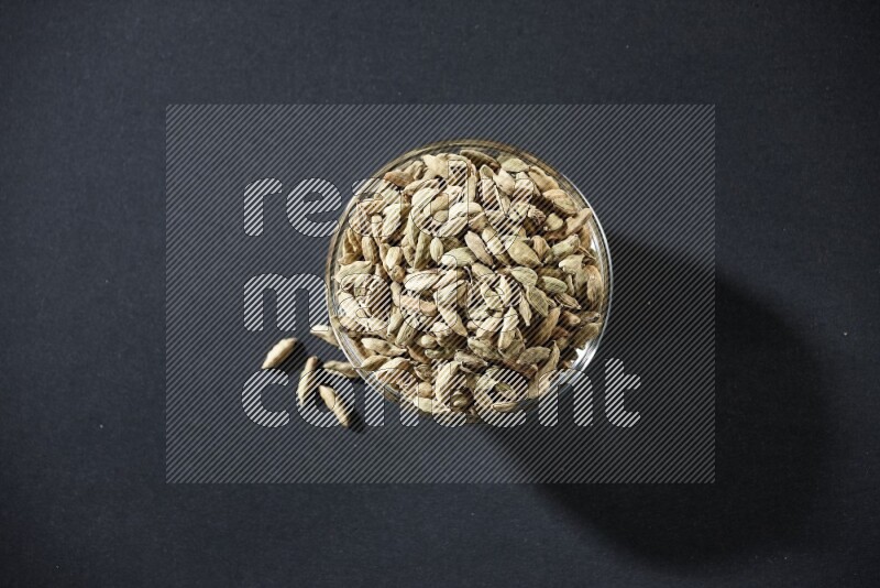 A glass bowl full of cardamom and more seeds spreaded beneath the bowl on black flooring
