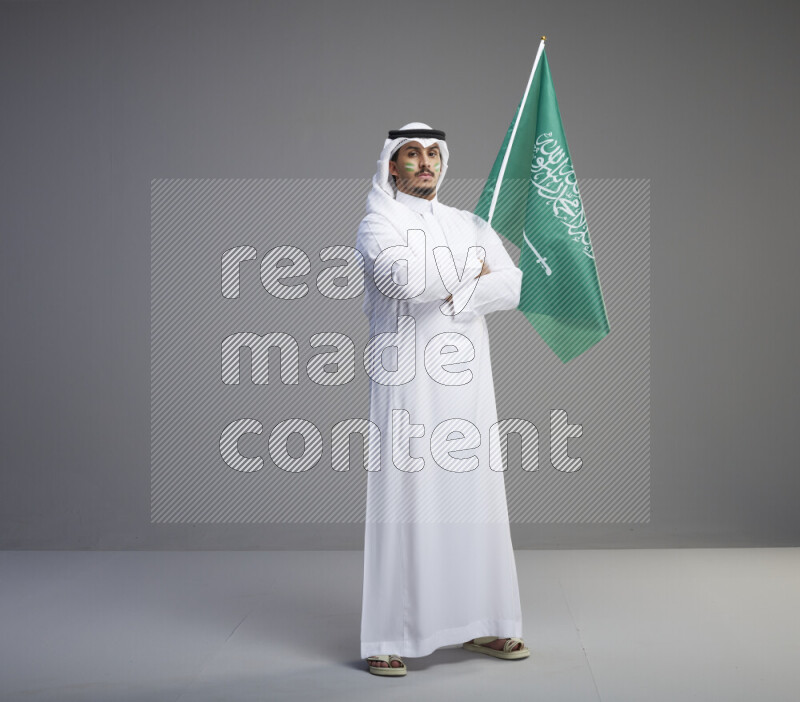 A Saudi man standing wearing thob and white shomag with face painting raising big Saudi flag on gray background