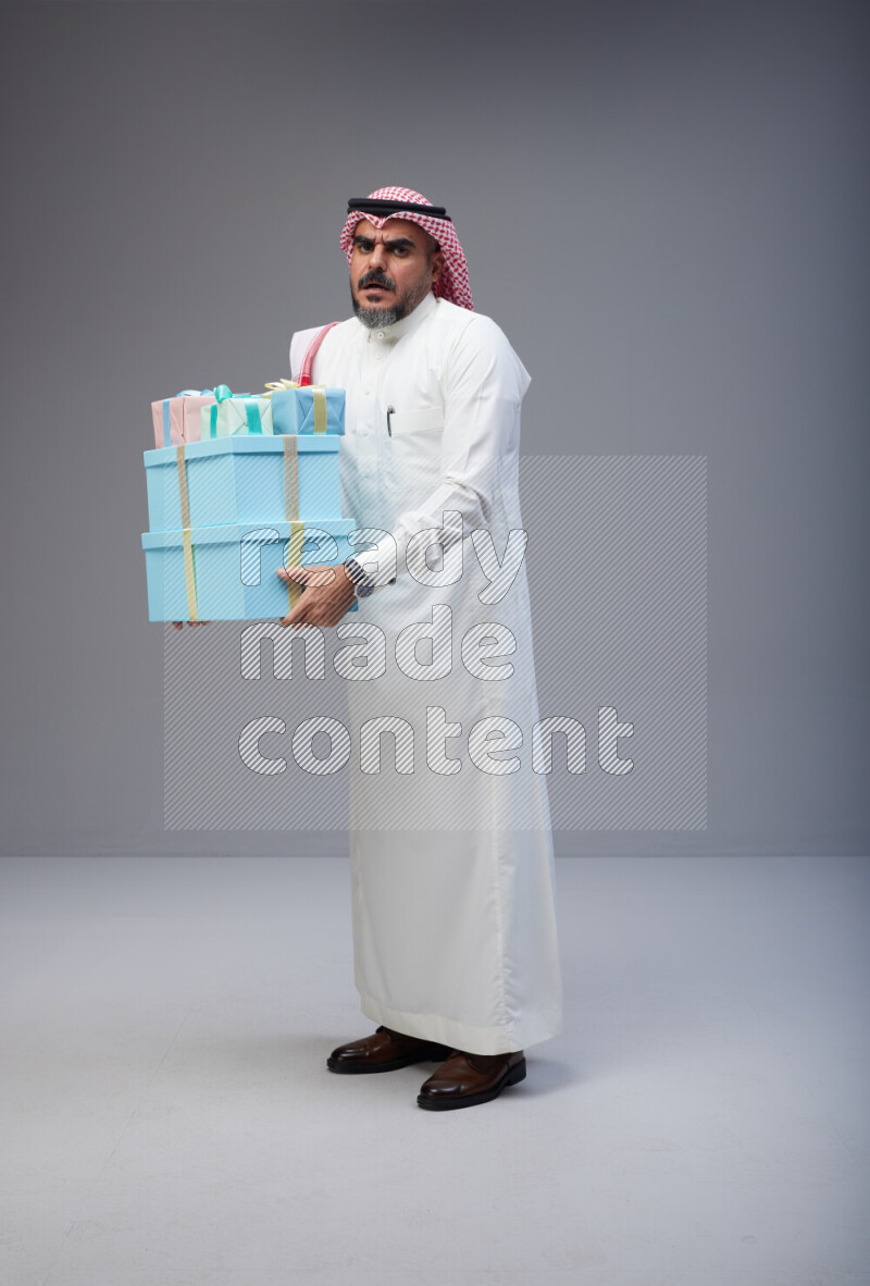 Saudi man Wearing Thob and red Shomag standing holding gift box on Gray background