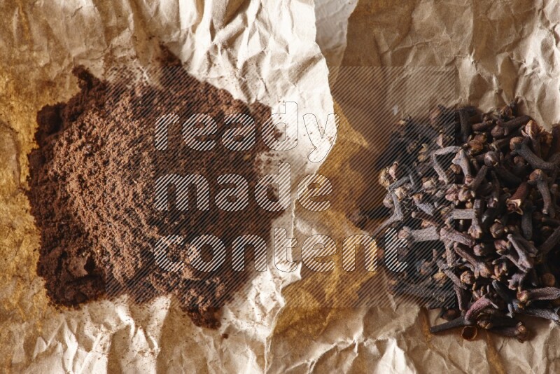 Cloves powder and cloves on 2 crumpled pieces of paper on a textured white flooring