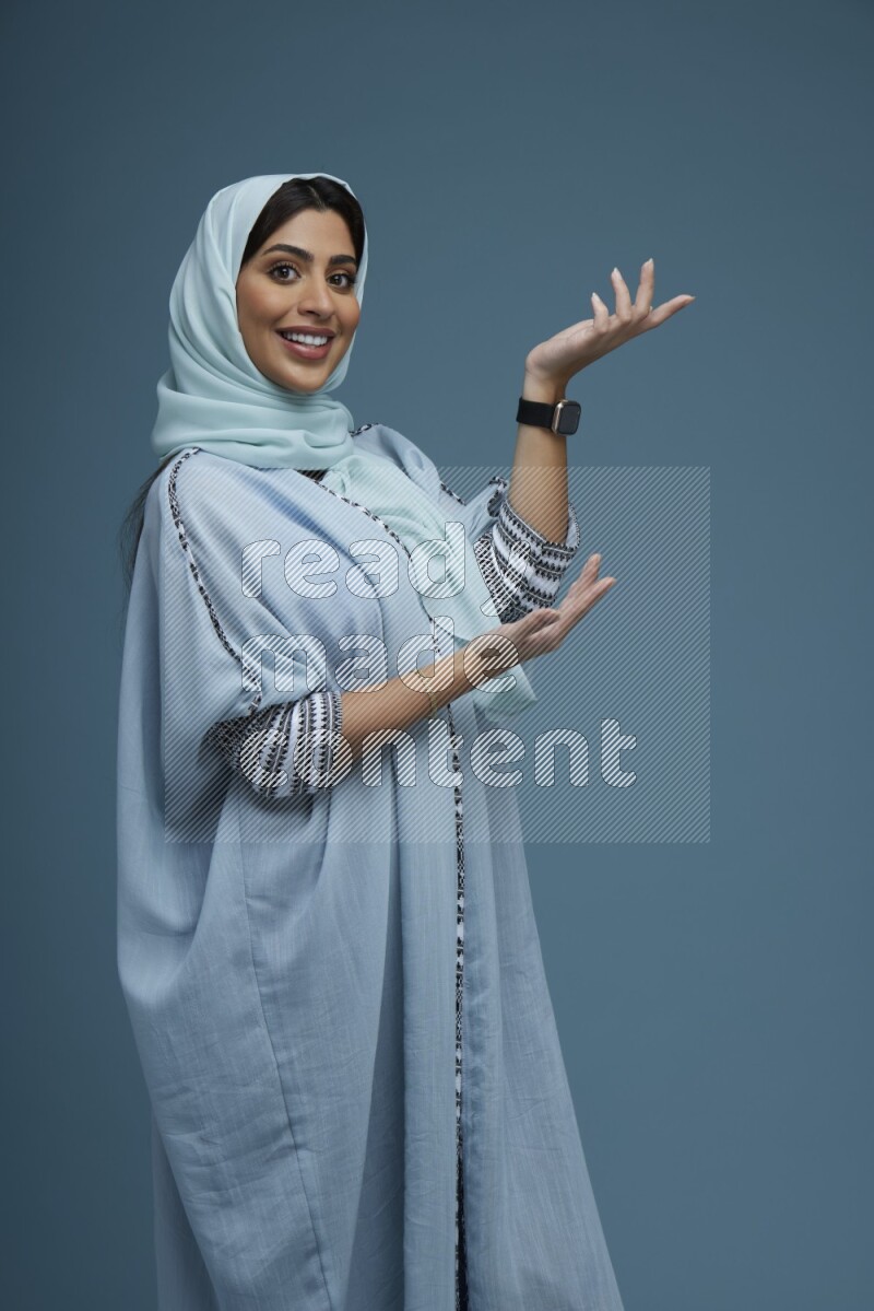 A Saudi woman pointing in a blue background wearing a Blue abaya