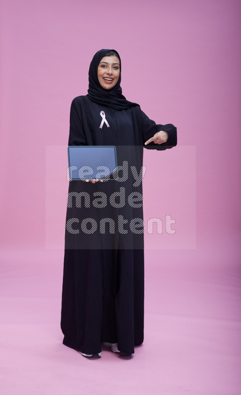Saudi woman wearing pink ribbon on Abaya standing showing a tablet to camera on pink background