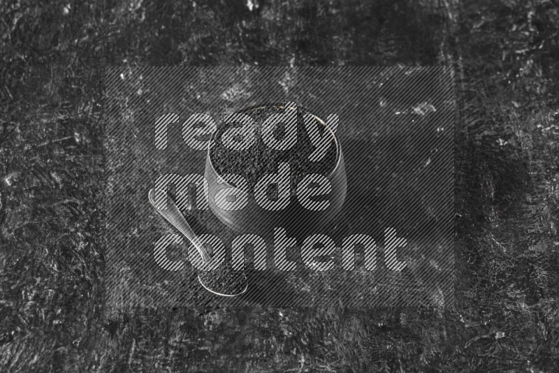 A black pottery bowl full of black seeds with a metal spoon full of the seeds on a textured black flooring