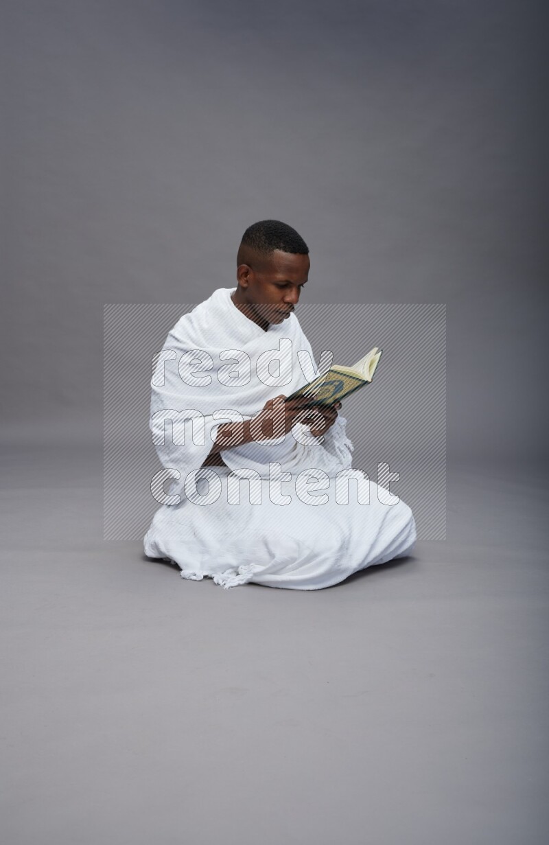 A man wearing Ehram sitting on floor reading quran on gray background