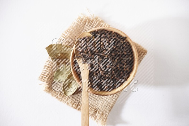 A wooden bowl and a wooden spoon full of cloves on a piece of burlap on a white flooring