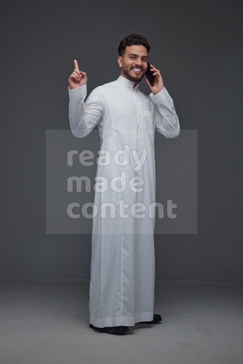 A Saudi man wearing Thobe and talking in the phone while standing and making different hand gestures eye level on a gray background