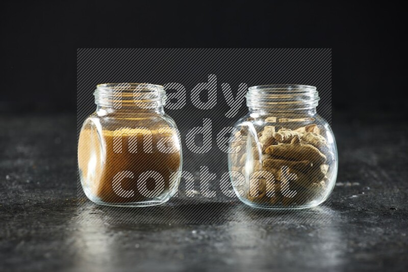 2 glass spice jars full of turmeric powder and dried whole fingers on textured black flooring