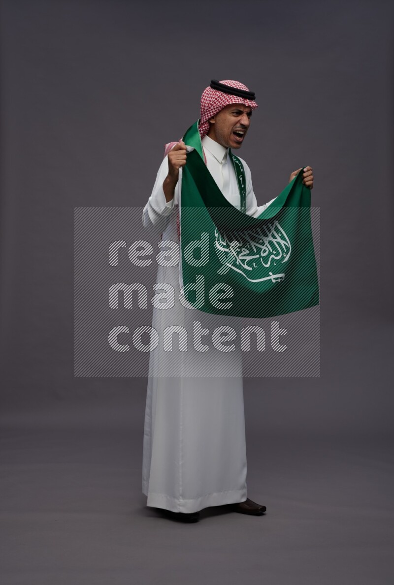 Saudi man wearing thob and shomag standing holding Saudi flag on gray background