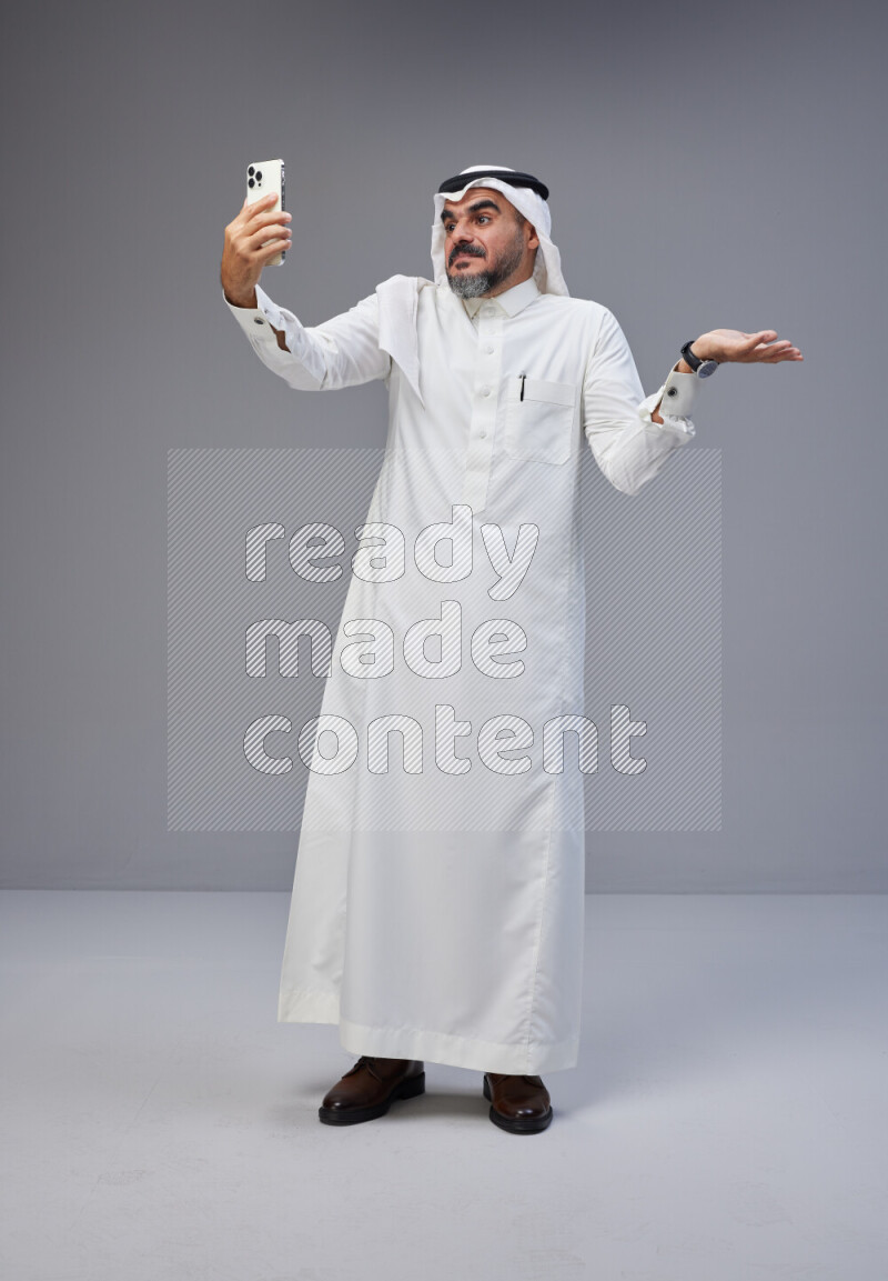 Saudi man Wearing Thob and white Shomag standing taking selfie on Gray background