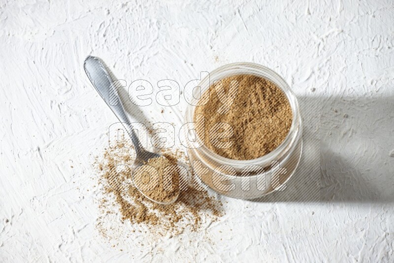 A glass jar and a metal spoon full of cumin powder on textured white flooring