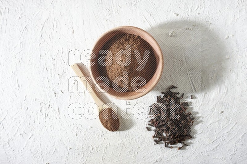 A wooden bowl and wooden spoon full of cloves powder with cloves spread on textured white flooring