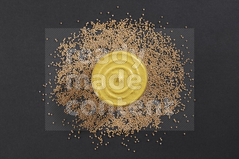 A glass bowl full of mustard paste with mustard seeds underneath on black flooring