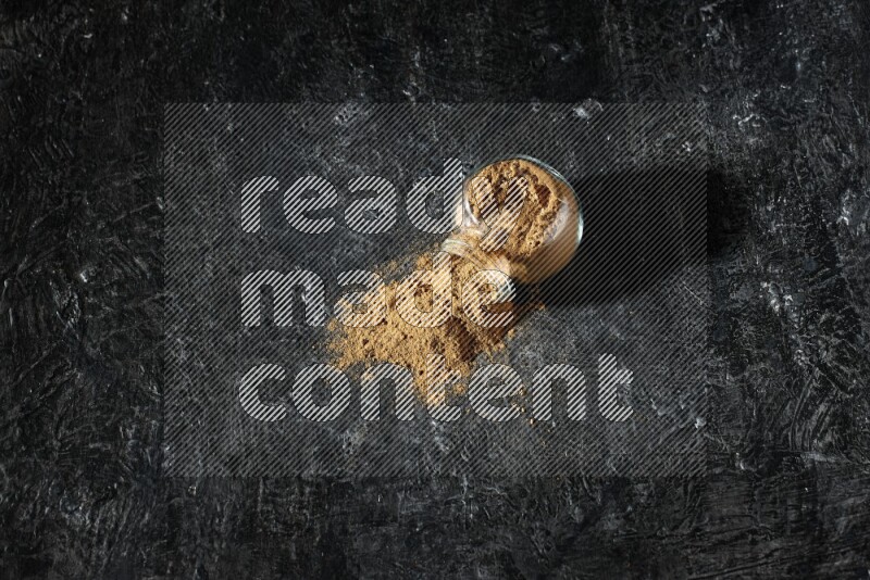 A flipped glass spice jar full of allspice powder and powder spilled out of it on a textured black flooring