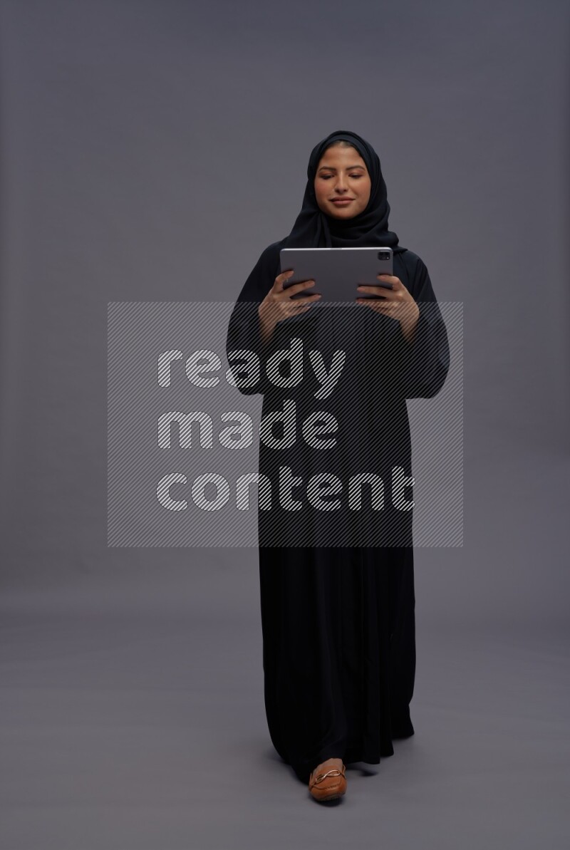 Saudi woman wearing Abaya standing working on tablet on gray background