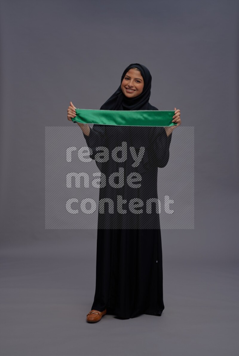 Saudi woman wearing Abaya standing holding Saudi flag on gray background