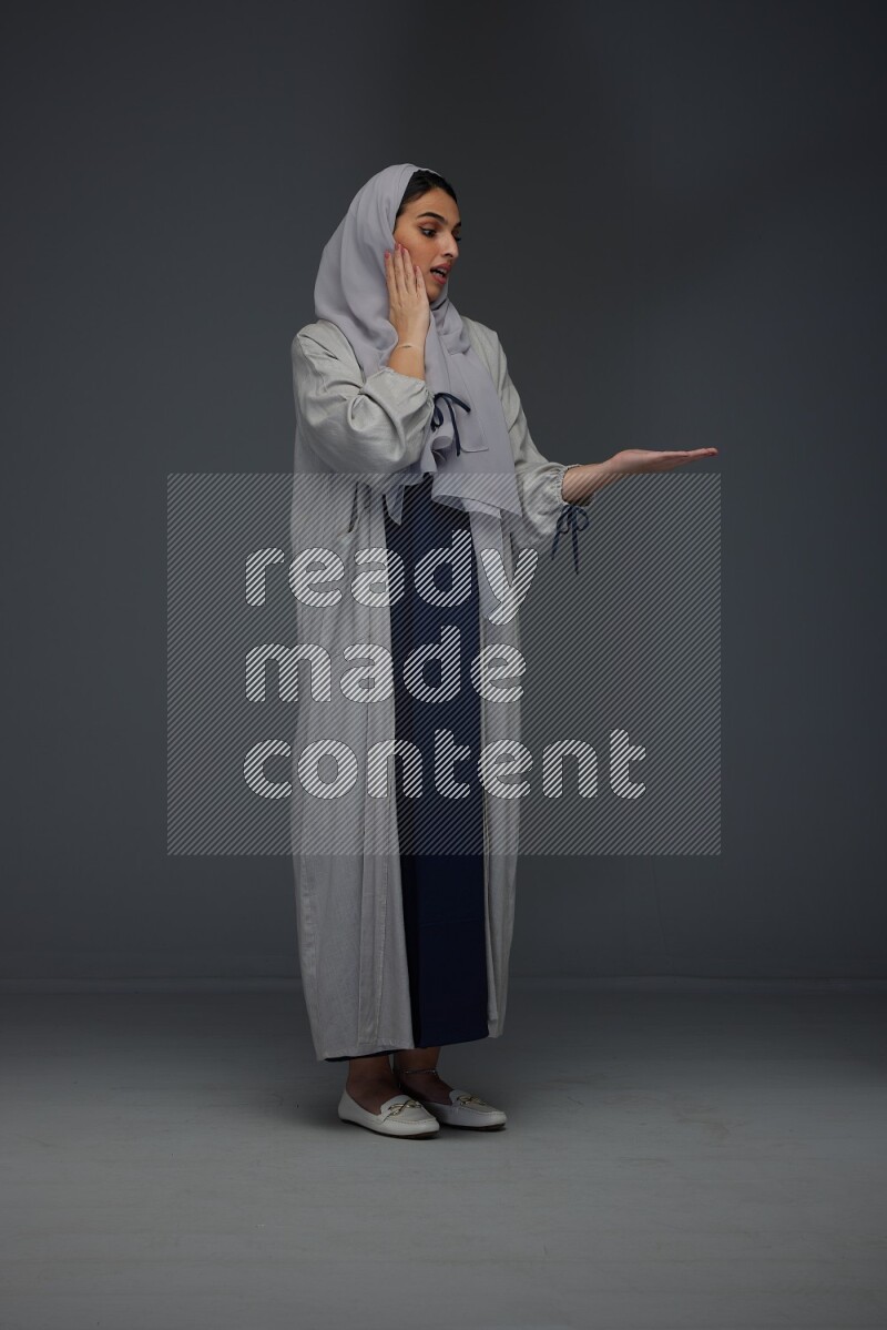A Saudi woman wearing a light gray Abaya and head scarf standing and pointing in different directions eye level on a grey background
