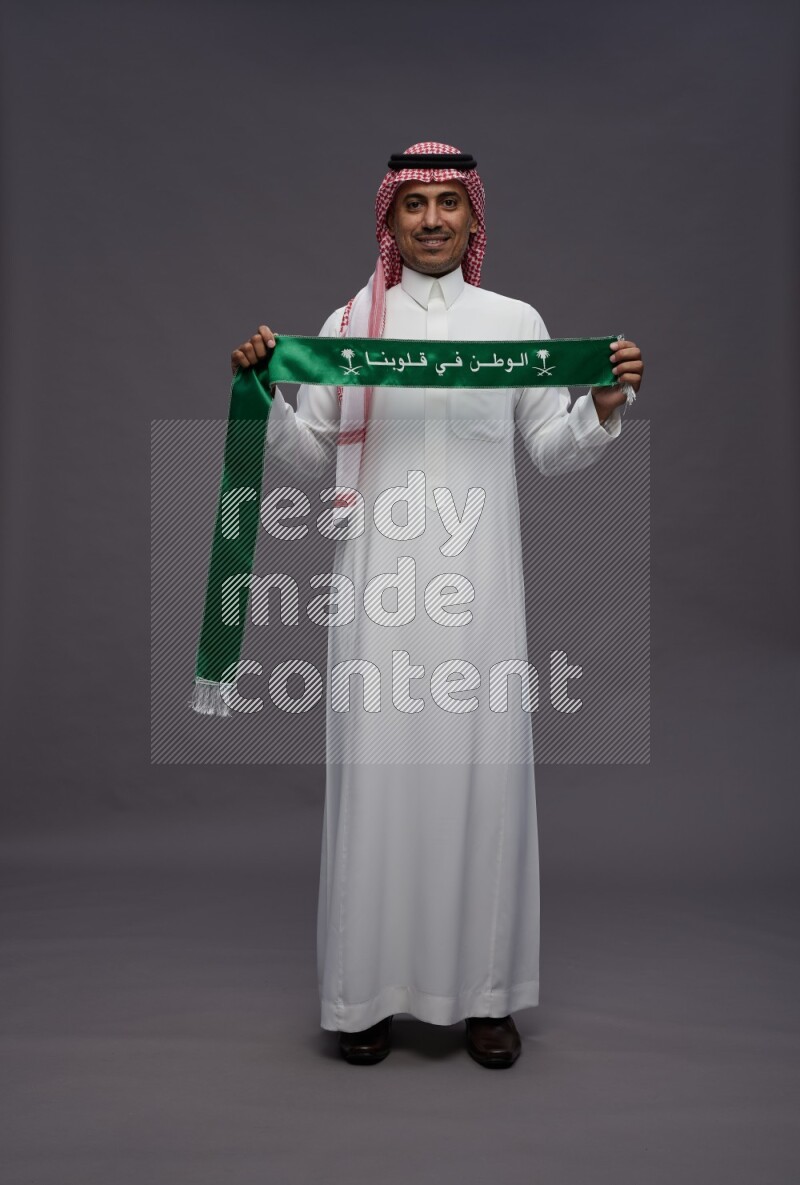 Saudi man wearing thob and shomag standing holding Saudi flag on gray background