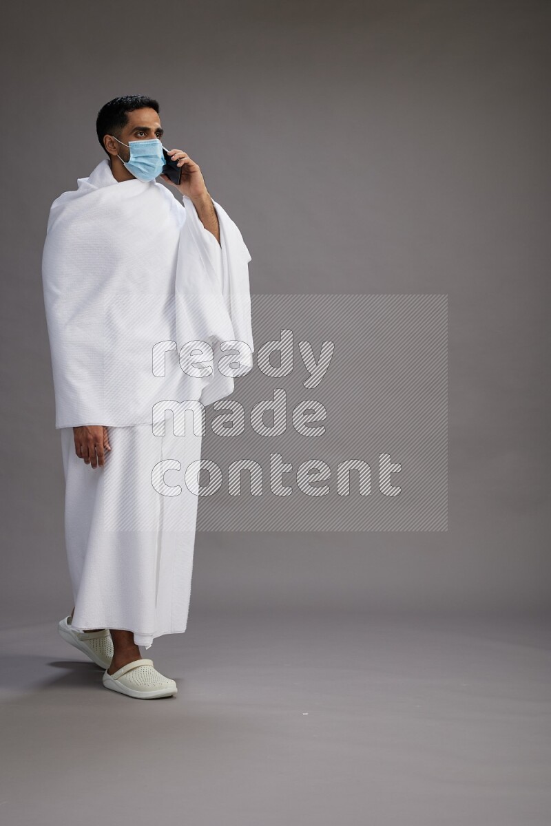 A man wearing Ehram with face mask Standing talking on phone on gray background