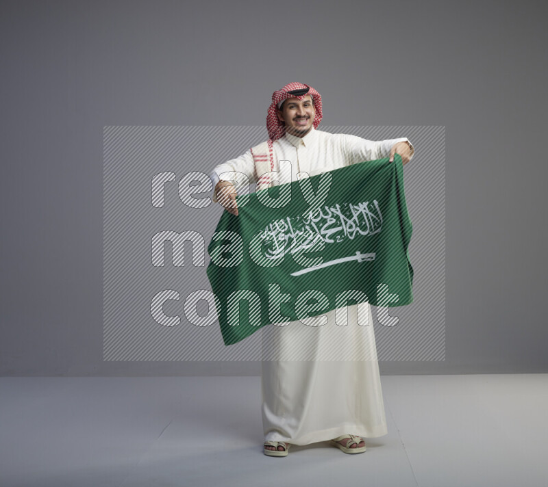 A saudi man standing wearing thob and red shomag holding big saudi flag on gray background