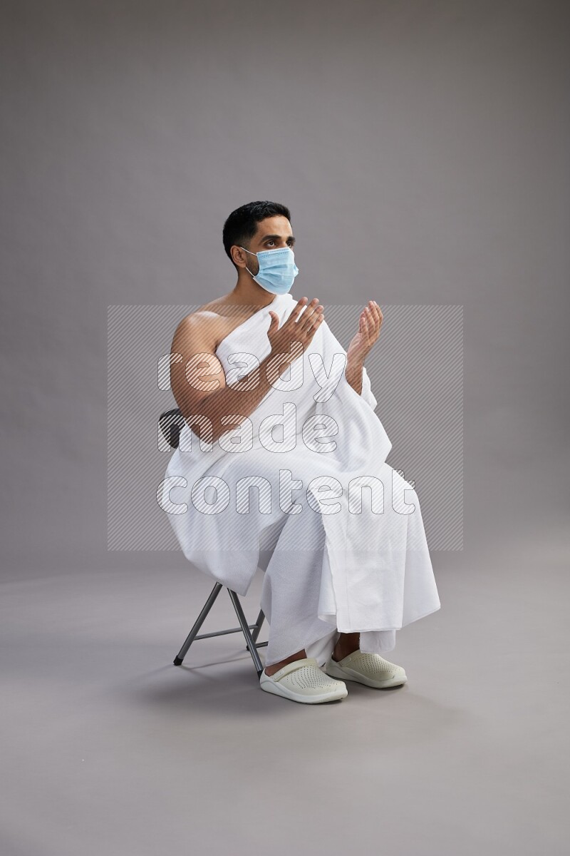 A man wearing Ehram with face mask sitting on chair dua'a on gray background