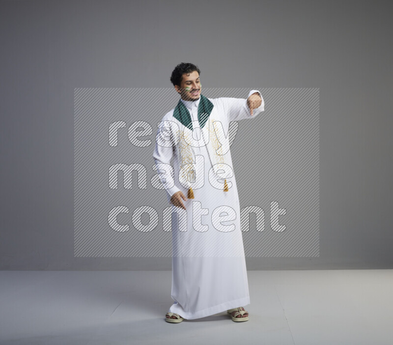 A Saudi man standing wearing thob and Saudi flag scarf with face painting on gray background