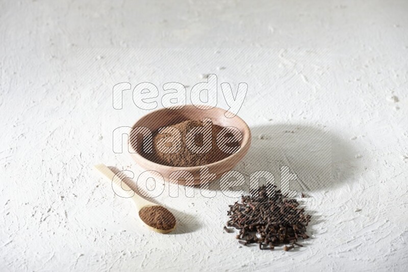 A wooden bowl and wooden spoon full of cloves powder with cloves spread on textured white flooring
