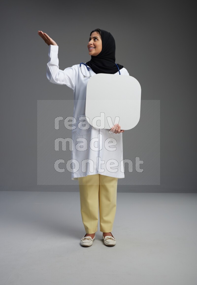 Saudi woman wearing lab coat with stethoscope standing holding social media sign on Gray background