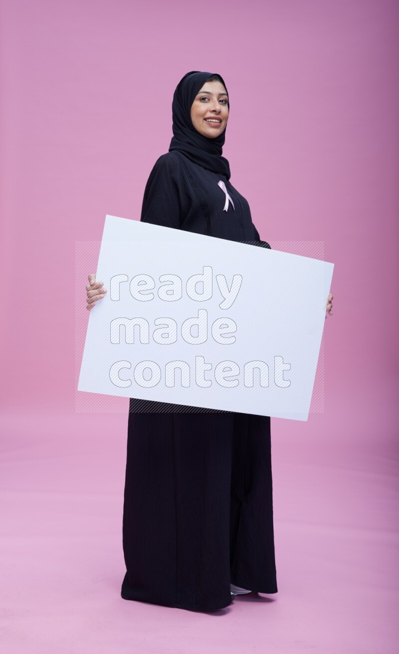 Saudi woman wearing pink ribbon on Abaya standing holding board on pink background