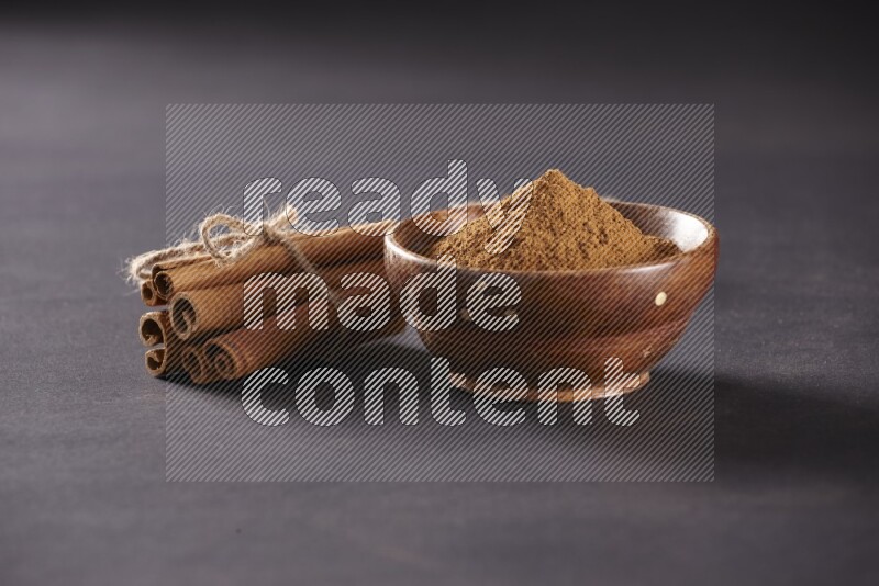 Cinnamon sticks stacked and bounded beside a wooden bowl full of cinnamon powder and a wooden spoon full of powder on black background