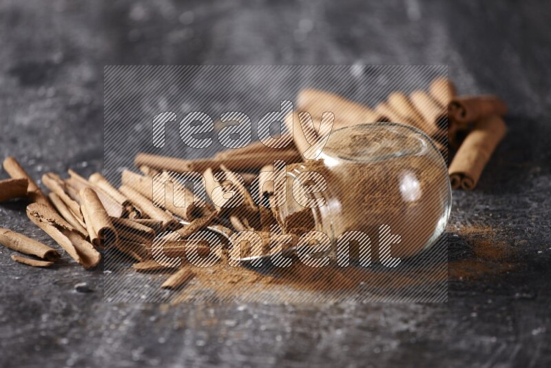 Herbal glass jar full cinnamon powder flipped and a metal spoon full of powder surrounded by cinnamon sticks on textured black background in different angles