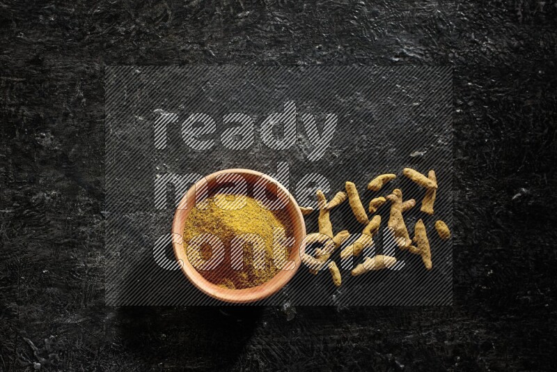 A wooden bowl full of turmeric powder with dried turmeric fingers on textured black flooring