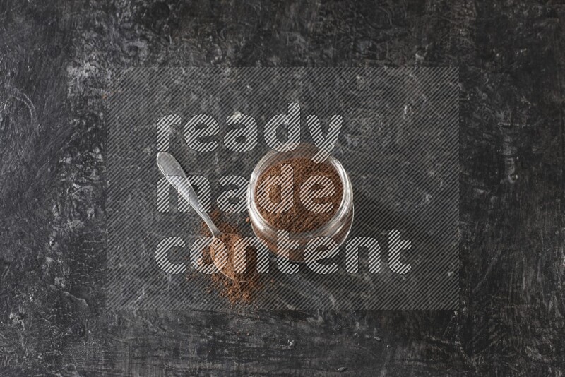 A glass jar full of cloves powder with a metal spoon on a textured black flooring