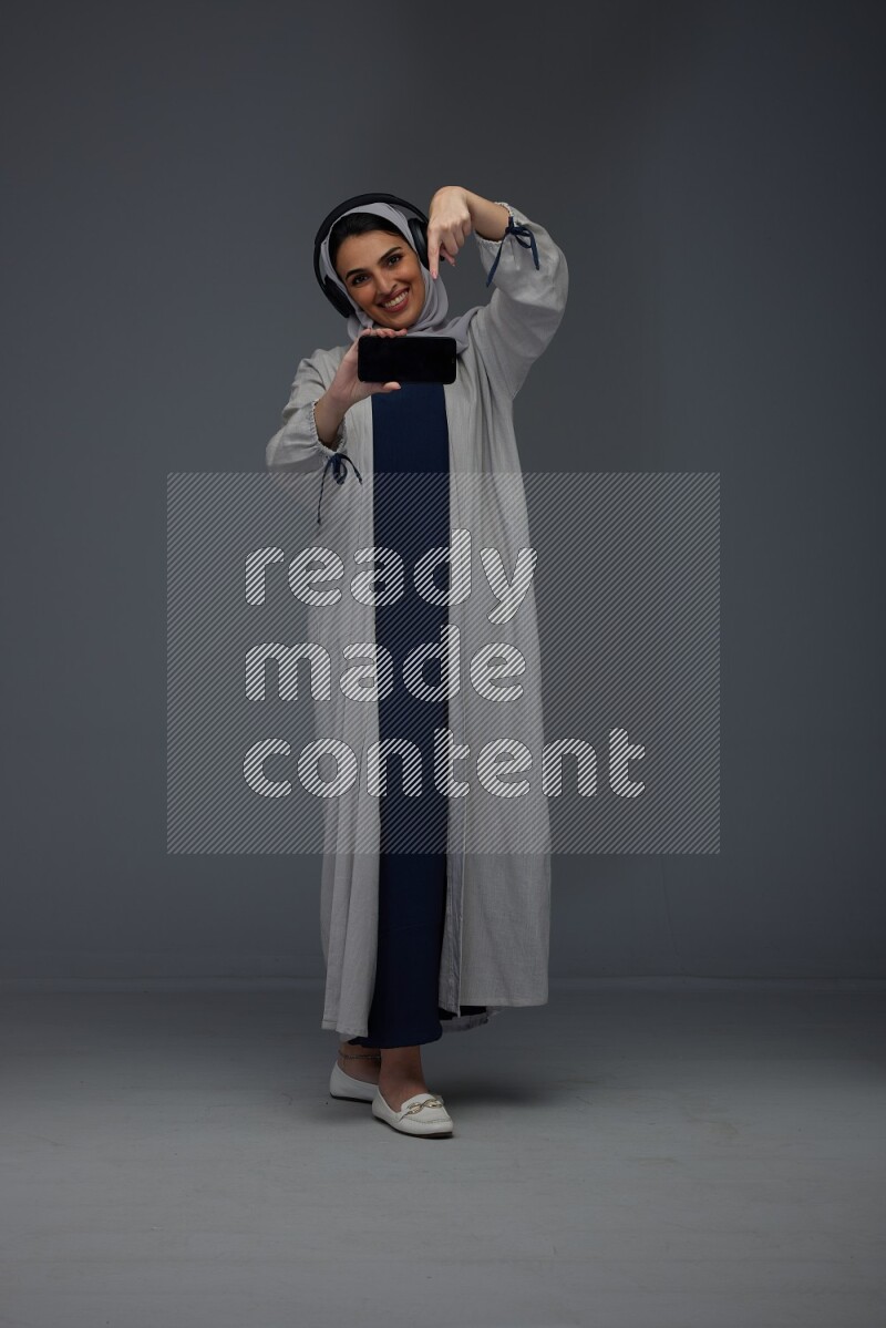 A Saudi woman wearing a light gray Abaya and head scarf setting on a dark grey chair and watching on the tablet eye level on a grey background