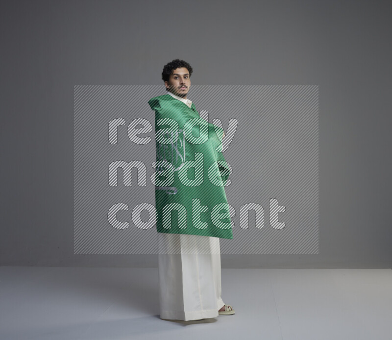 A Saudi man standing wearing thob wrapping big Saudi flag on gray background