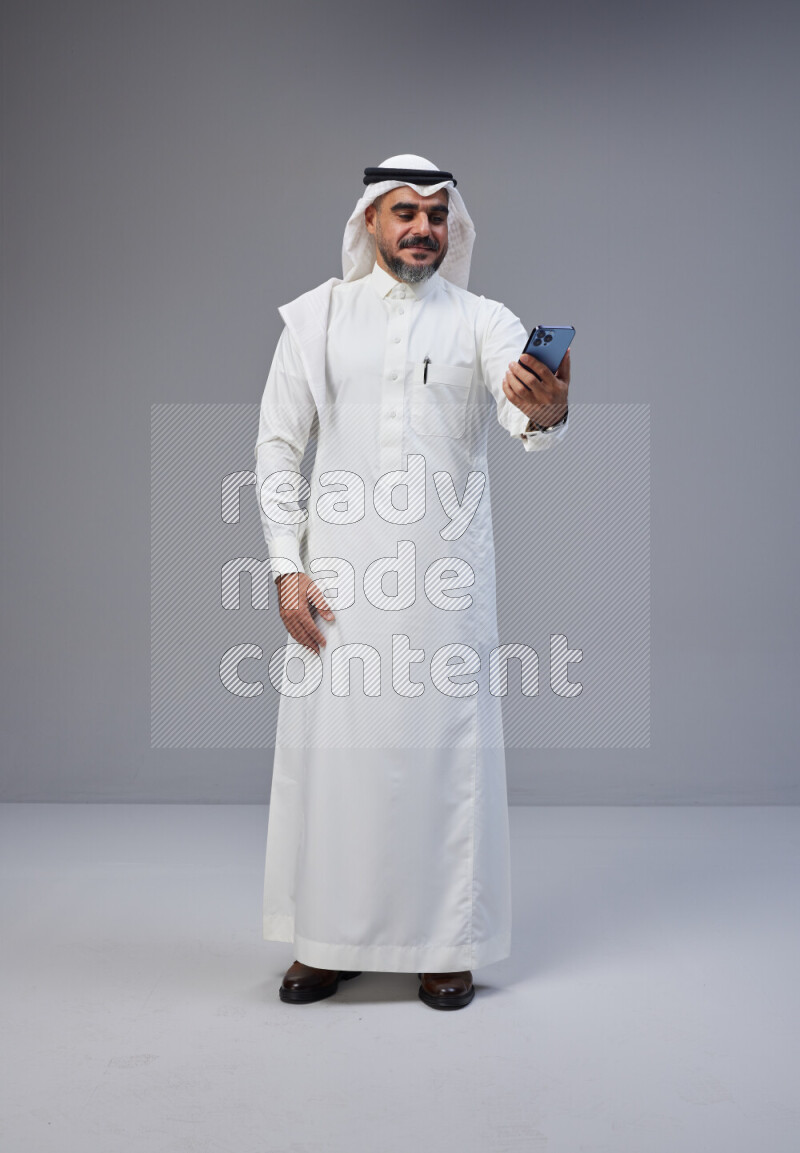 Saudi man Wearing Thob and white Shomag standing taking selfie on Gray background