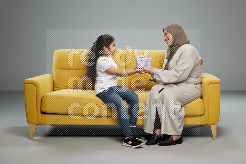A girl sitting on a yellow sofa and giving a gift to her mother on gray background