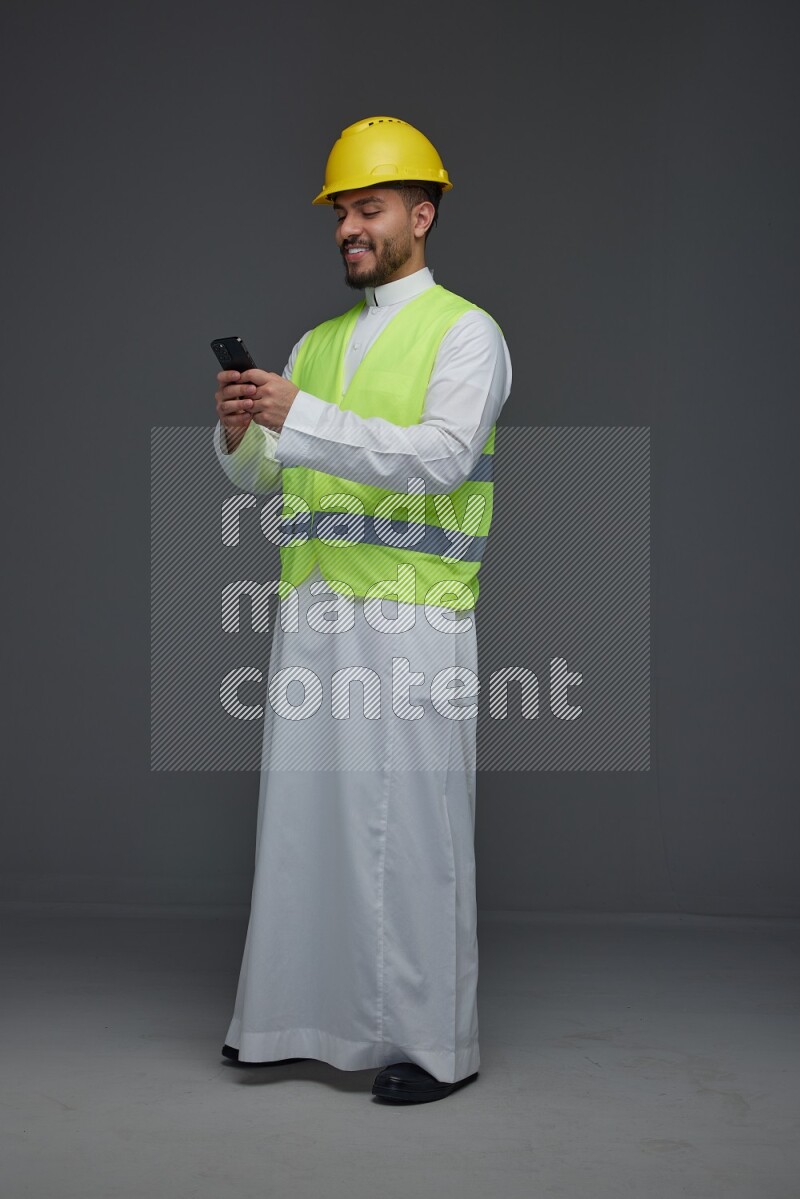 A Saudi man wearing Thobe with a yellow safety vest and white helmet standing and using his phone eye level on a gray background