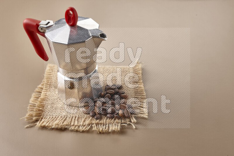 A moka pot with red handle surrounded by roasted coffee beans on beige background