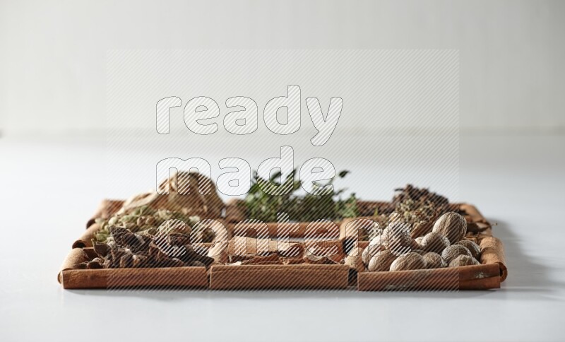 9 squares of cinnamon sticks, the middle square is empty and surrounded with dried mint, dried ginger, cardamom, star anise, cinnamon, nutmeg, dried basil and cloves on white flooring