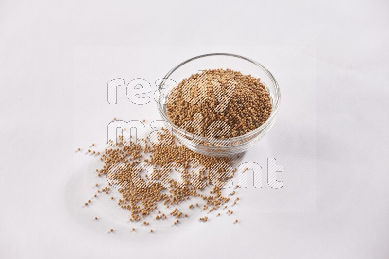 A glass bowl full of mustard seeds and more seeds spread on a white flooring