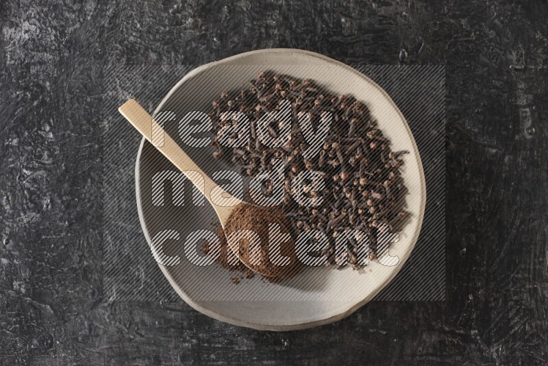 A Pottery plate full of cloves and a wooden spoon full of cloves powder on it on a textured black background