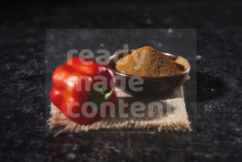 A wooden bowl full of ground paprika powder with a red bell pepper on a burlap fabric on black background