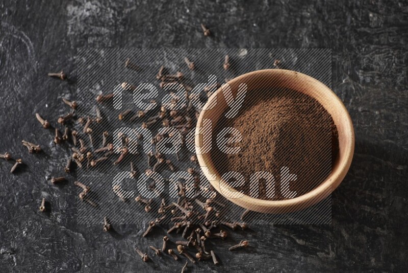 A wooden bowl full of cloves powder on a textured black flooring