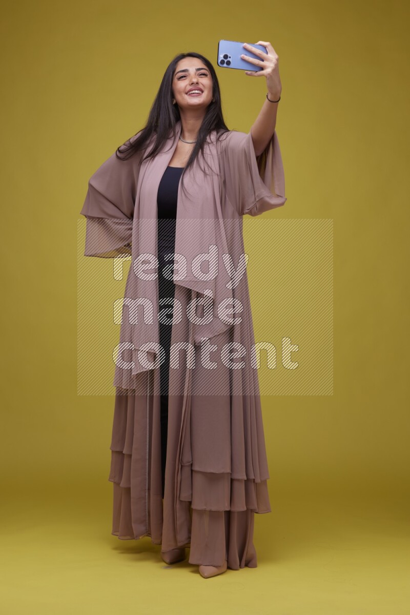A woman Taking a Selfie on a Yellow Background wearing Brown Abaya