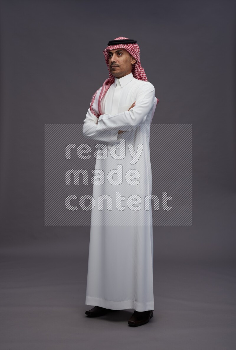 Saudi man wearing thob and shomag standing with crossed arms on gray background