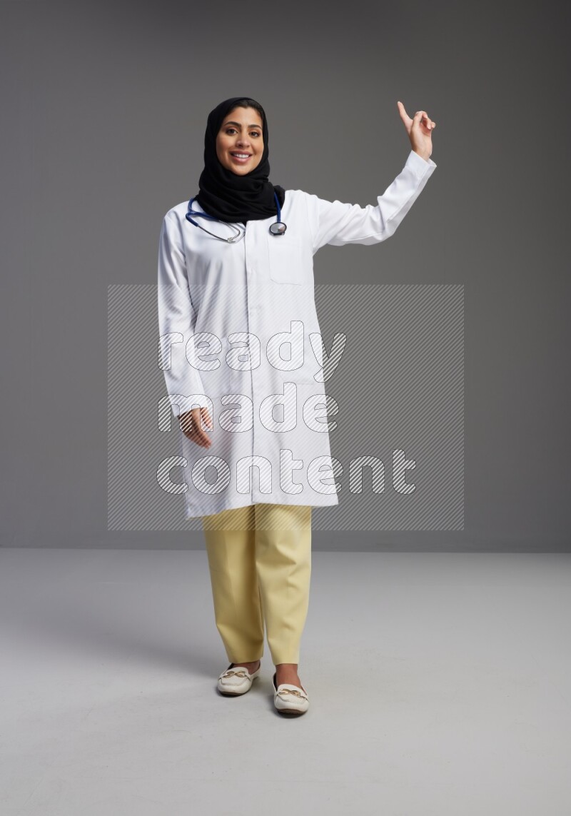 Saudi woman wearing lab coat with stethoscope standing interacting with the camera on Gray background