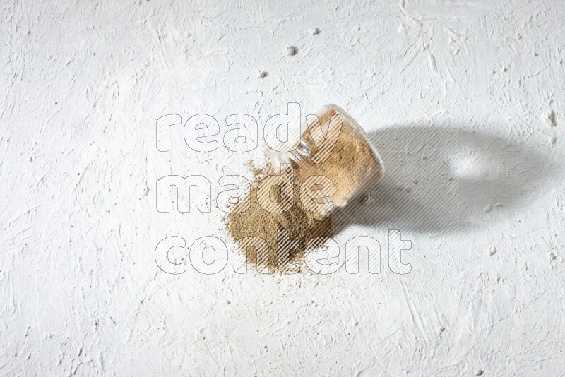 A flipped glass jar full of cumin powder and powder spilled out on textured white flooring