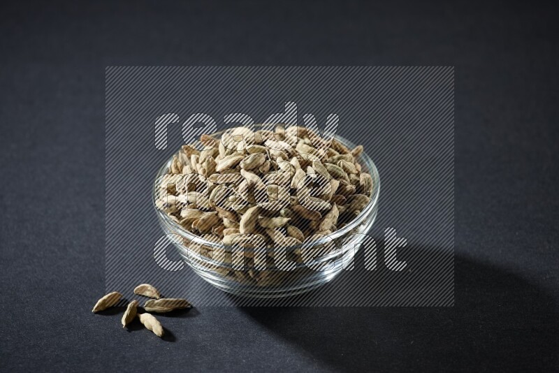 A glass bowl full of cardamom and more seeds spreaded beneath the bowl on black flooring