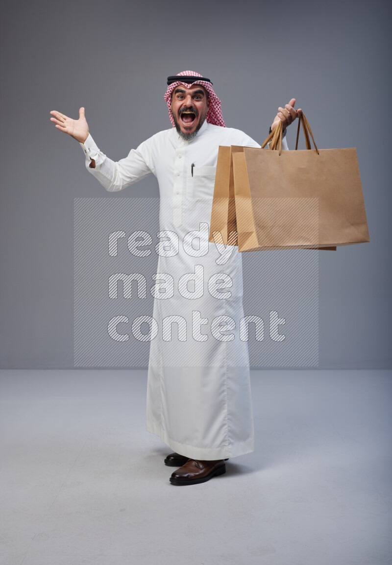 Saudi man Wearing Thob and red Shomag standing holding shopping bag on Gray background