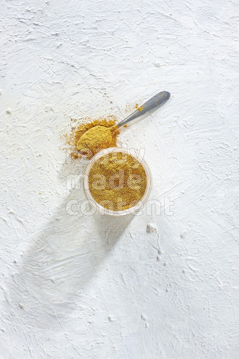A glass jar and a metal spoon full of turmeric powder on a textured white flooring
